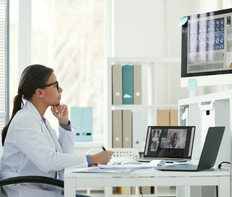 Young female doctor sitting at her workplace in front of computer monitor and listening to her colleagues during online conference