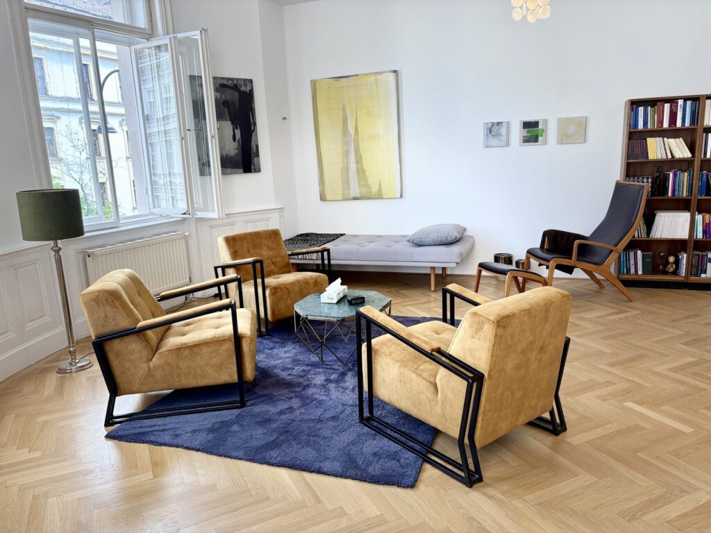 Heller Behandlungsraum der Praxis Las Almas in Wien mit Sesselgruppe, Bücherregal und Parkettboden