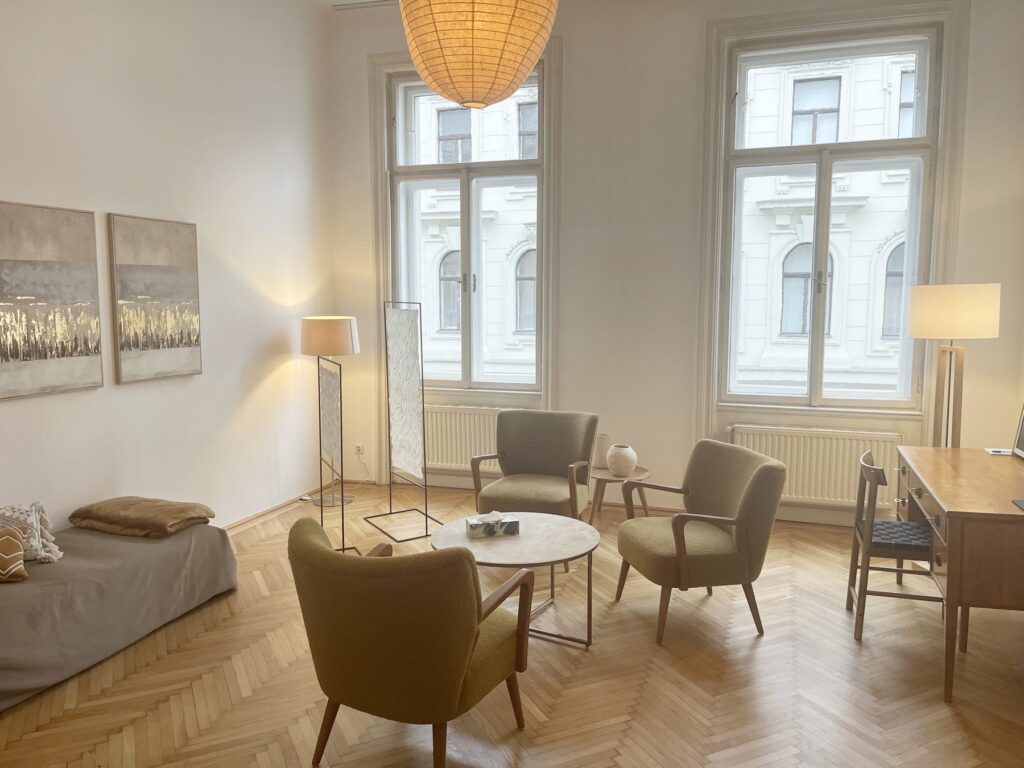 Gesprächsraum der Praxis Las Almas in Wien mit Sesseln, Liege und Schreibtisch vor hohen Altbaufenstern